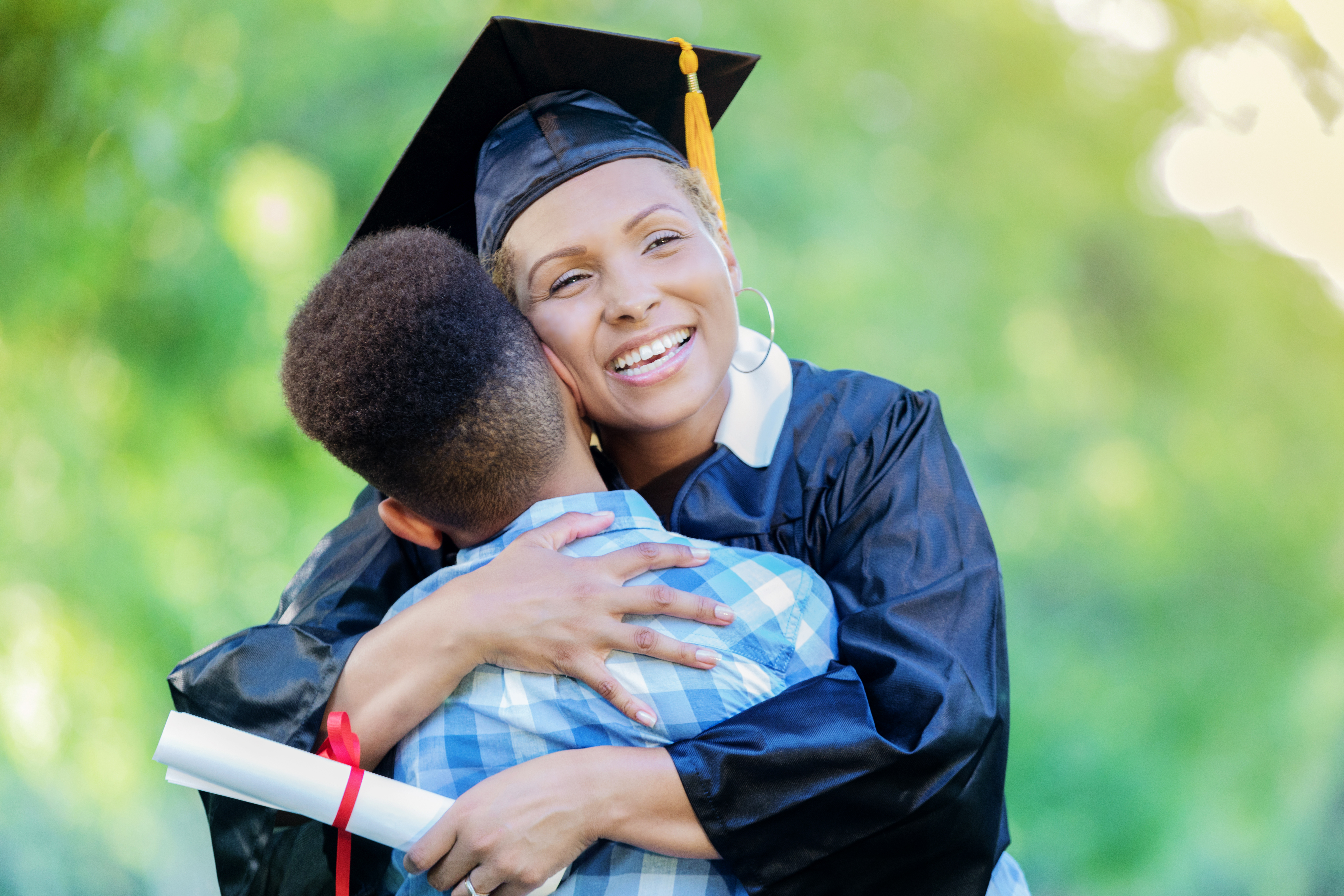 Smiling mba graduate in cap and gown hugs her young son while holding a diploma outdoors after the ceremony