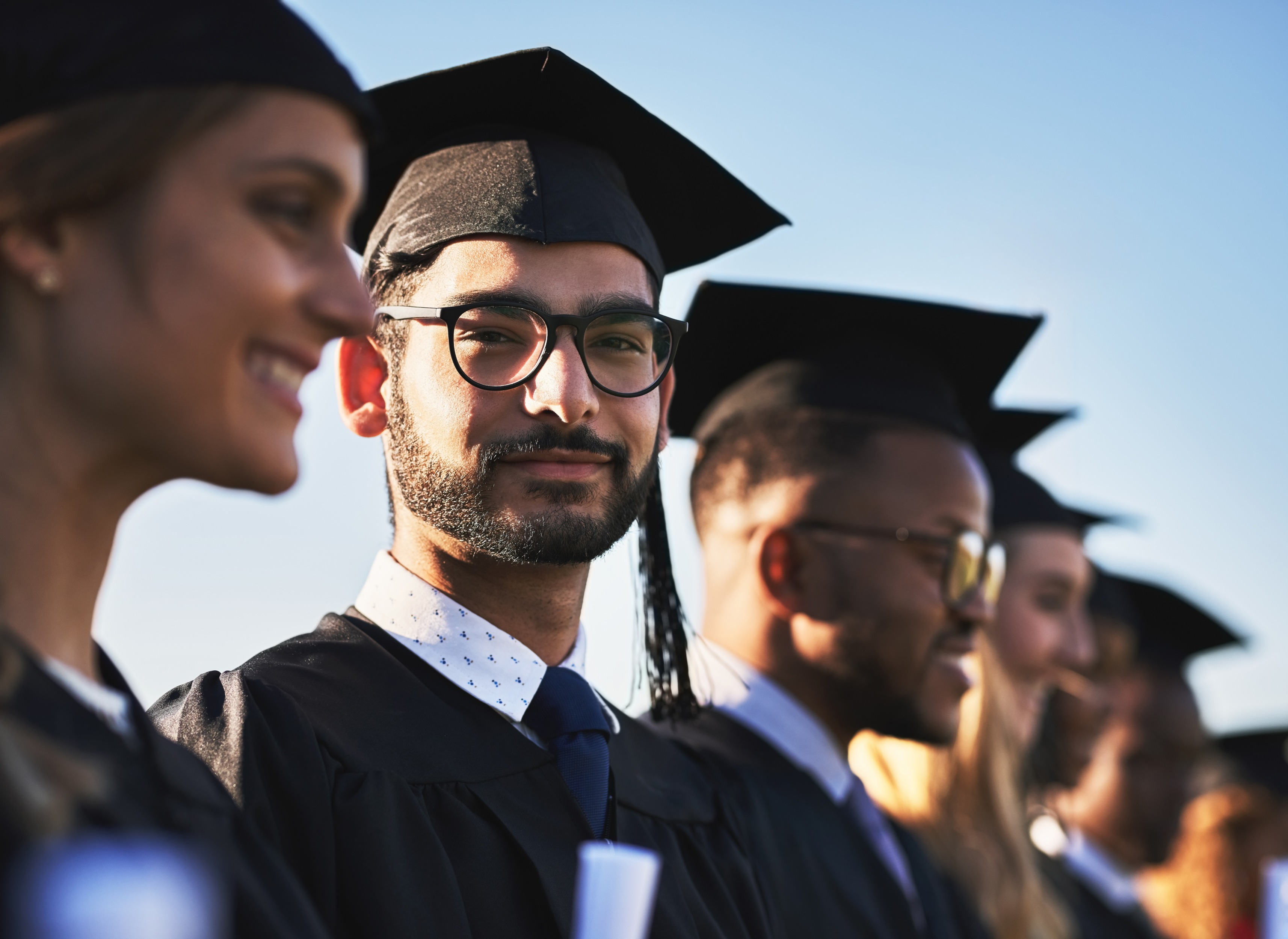 A masters degree student looks at camera with a smirk while standing among fellow graduates.