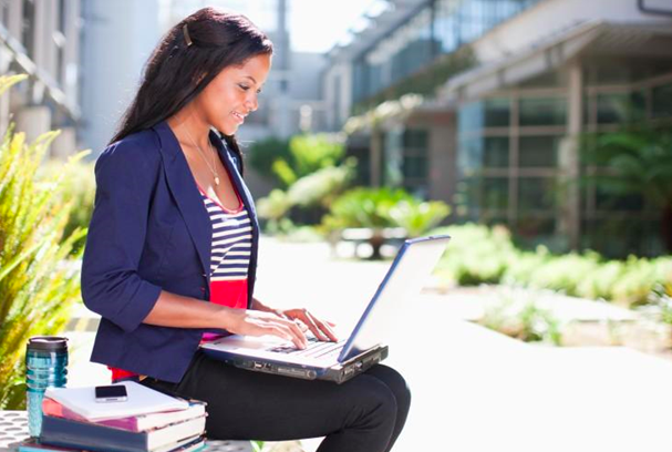 An online MBA student sitting on a bench outside and typing on a laptop