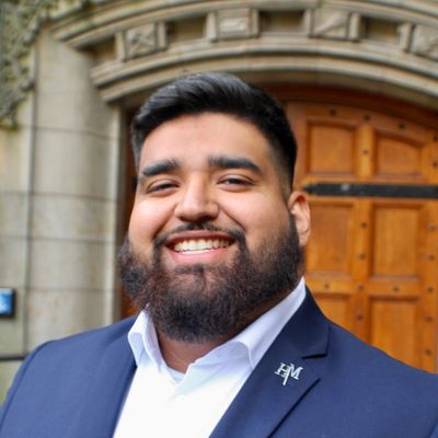 Marco Hernandez, the NDNU Career Services and Campus Engagement Coordinator, smiling in a blue blazer and white shirt on the NDNU campus.