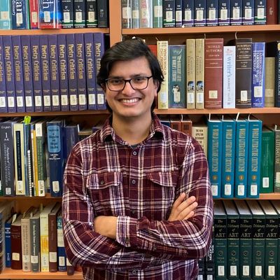 A student smiling with arms folded, standing in front of shelves filled with reference books.