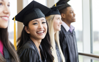 A smiling graduate wearing a cap and gown stands with other graduates.